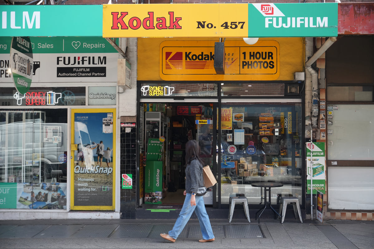A person walks past a vintage Kodak No. 457 instant photo camera store on a city sidewalk.